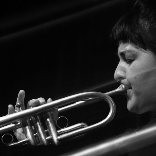 Black and white close-up photograph of a woman in profile playing a silver trumpet, with dramatic lighting against a dark background.