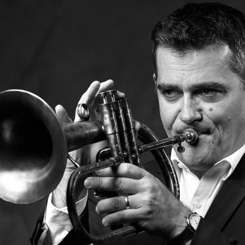 A black and white close-up photograph of a man in a dark suit playing a trumpet, with the trumpet bell prominently visible on the left side against a dark background.