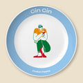 A round decorative plate with a white center, a light blue rim, and printed text reading 'Cin Cin' at the top and 'Italian Festa' at the bottom. The center features a stylized illustration of a person in an Italian flag color outfit holding a red rose.