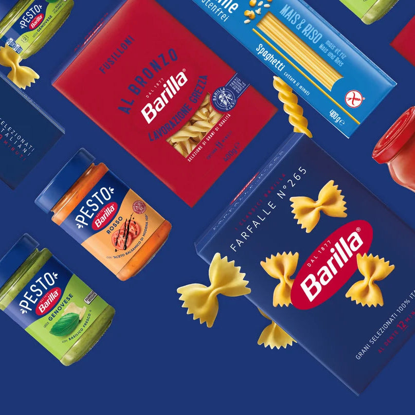 A collage of Barilla pasta products and pesto jars floating diagonally on a dark blue background, including red and blue boxes, yellow bowties, and colorful pesto containers.