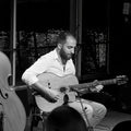 Three musicians performing live jazz on stage in a club setting, featuring two acoustic guitar players and an upright bassist, with a 'Jazz Club' sign visible in the background.