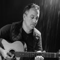 A man wearing a black quilted jacket and dark pants sits and plays an acoustic guitar under purple stage lighting against a dark curtain background.