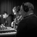 A black and white photograph of a musician in a recording studio, shown from behind wearing over-ear headphones and facing a keyboard synthesizer, with other band members blurred in the background.